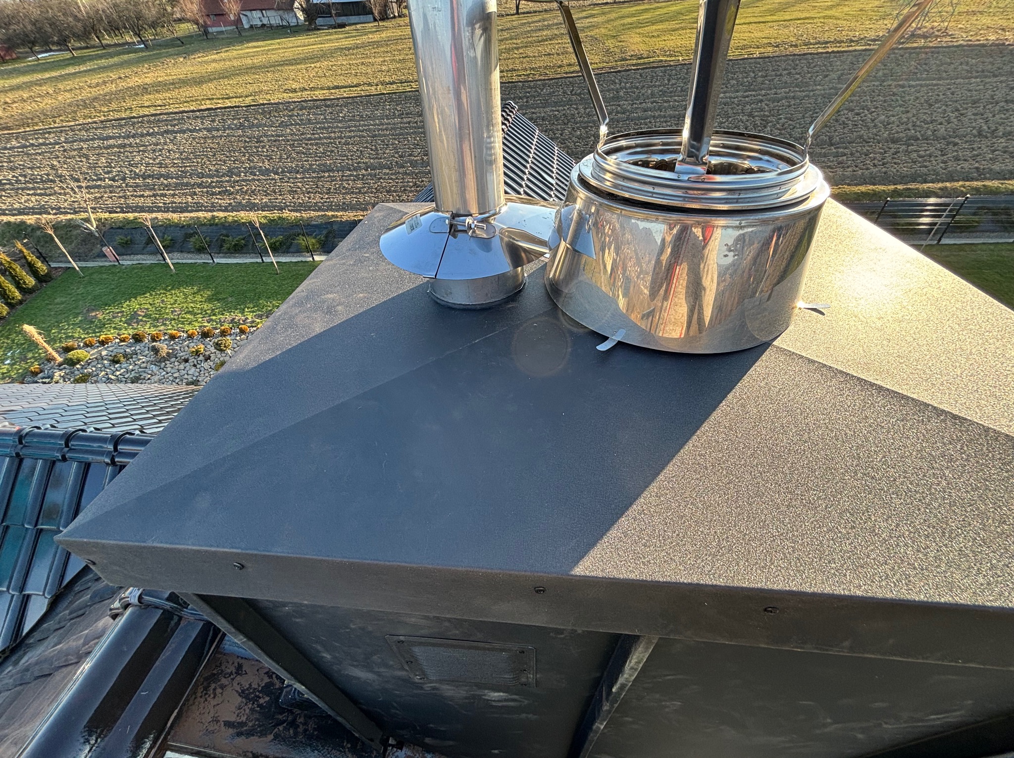 Metalowa czapka kominowa z dwoma przewodami wentylacyjnymi na tle pola i zieleni.