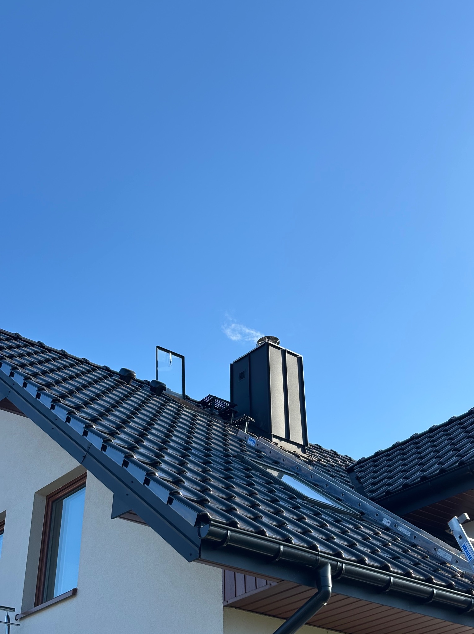 Czarna dachówka ceramiczna na skośnym dachu domu jednorodzinnego, widoczny komin, okno dachowe, rynna i drabina oparta o krawędź dachu na tle błękitnego nieba.