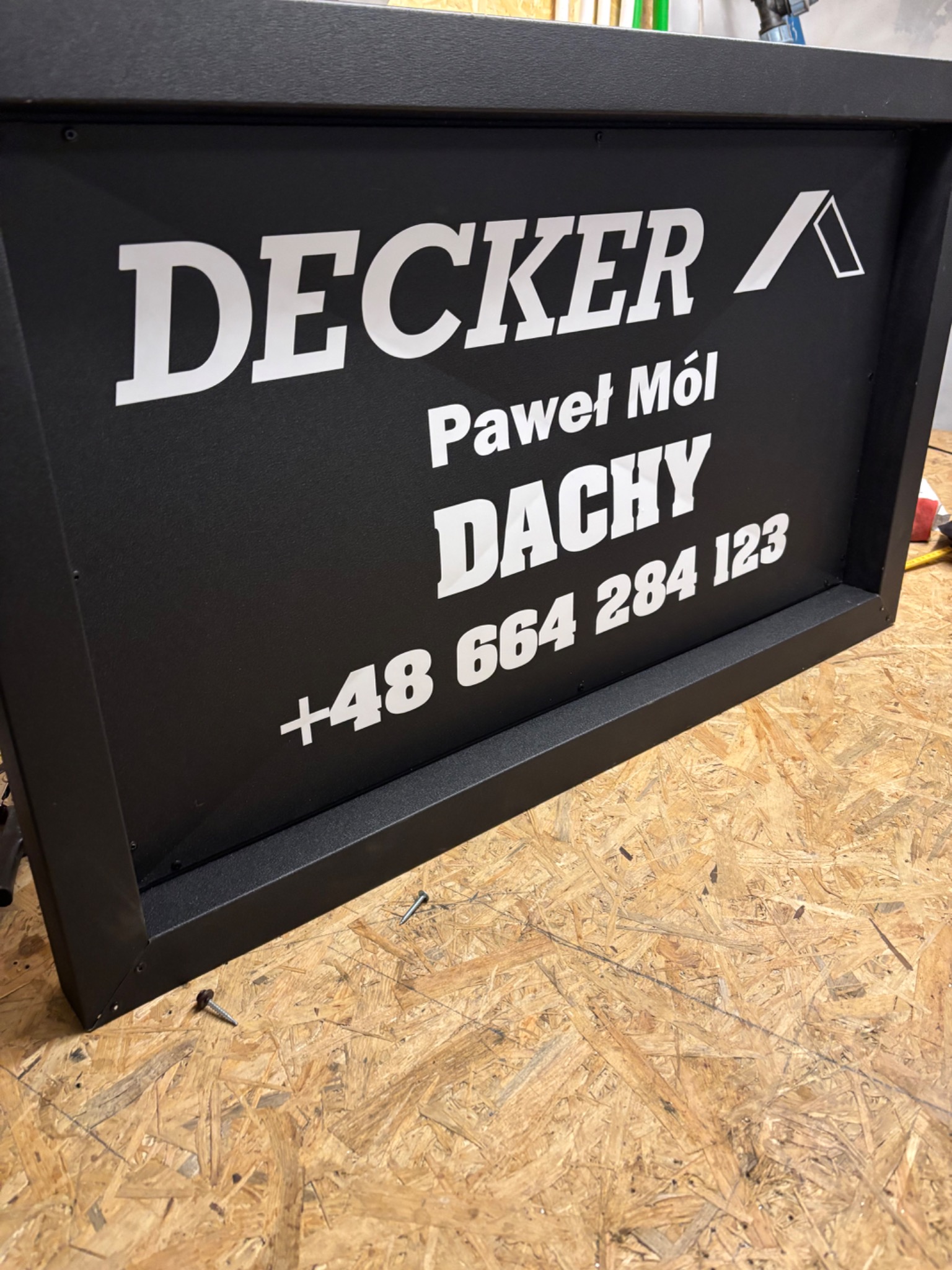 Czarna tablica reklamowa z białym napisem 'DECKER Paweł Mól DACHY +48 664 284 123' na tle płyty OSB.