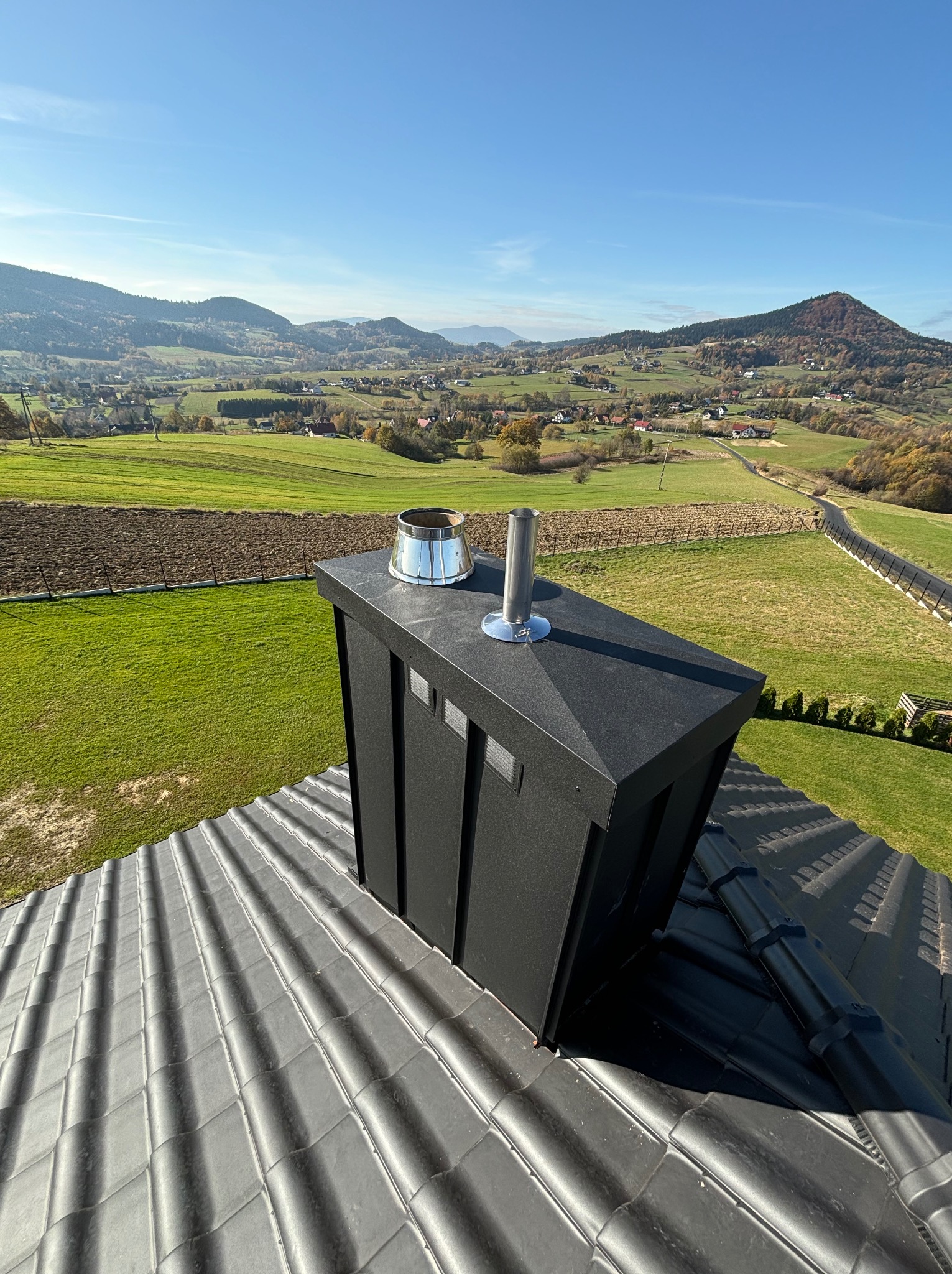 Czarny, nowoczesny komin z metalowym zakończeniem na dachu z ciemnej dachówki ceramicznej, w tle pagórkowaty krajobraz.