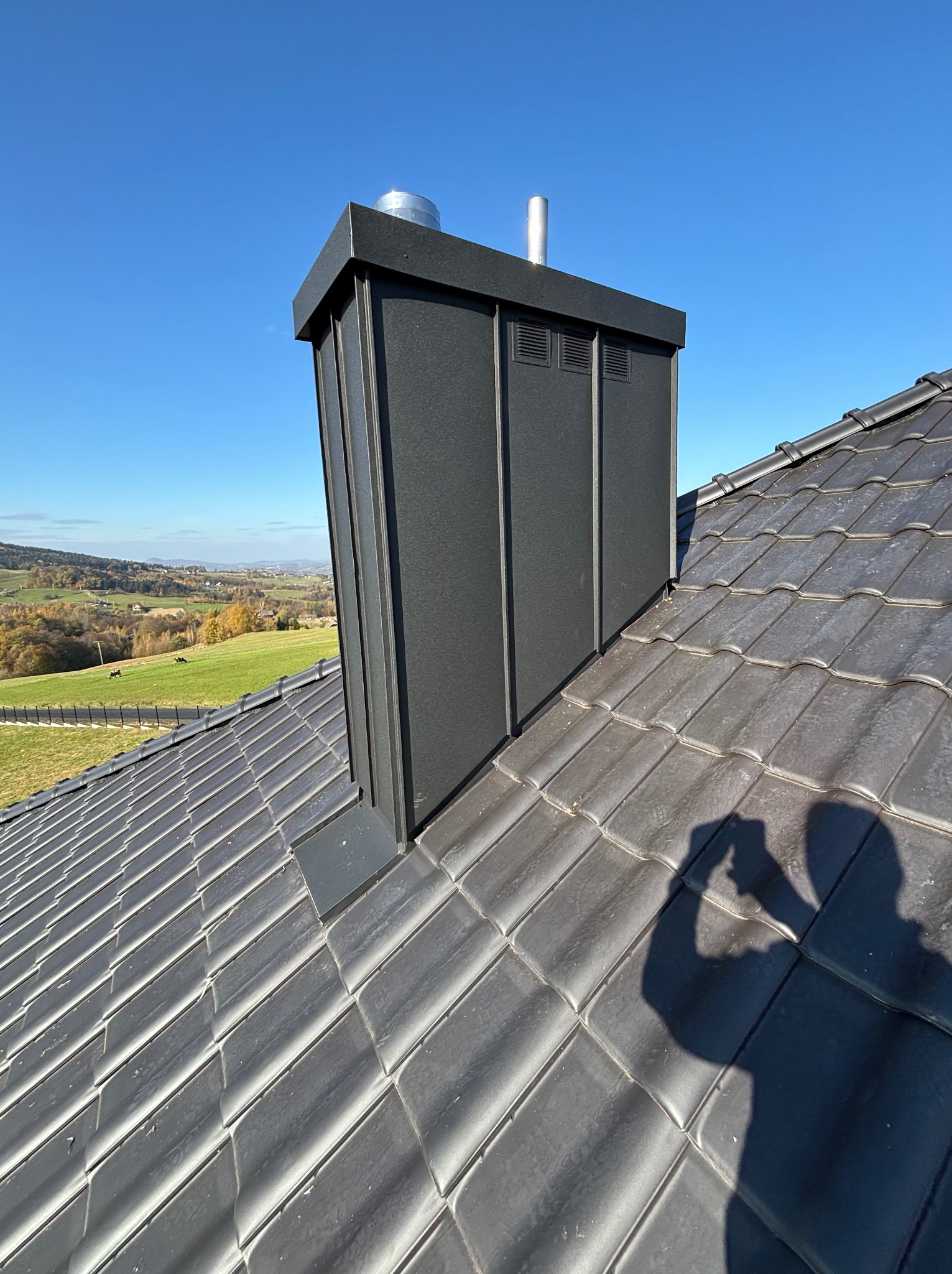 Nowoczesny komin obłożony czarną blachą na tle ceramicznej dachówki, widok na pagórkowaty krajobraz w słoneczny dzień.