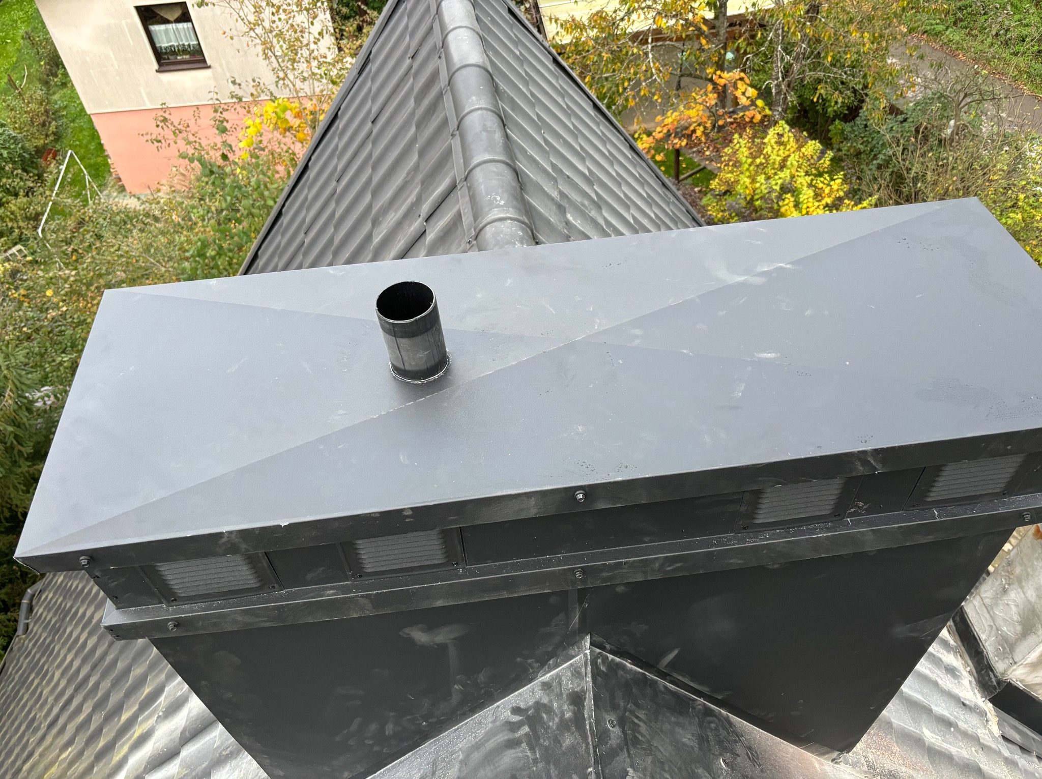 Szare pokrycie komina z krótką rurą wentylacyjną na szarym dachu z blachy, widok z góry na tle zieleni i budynku.