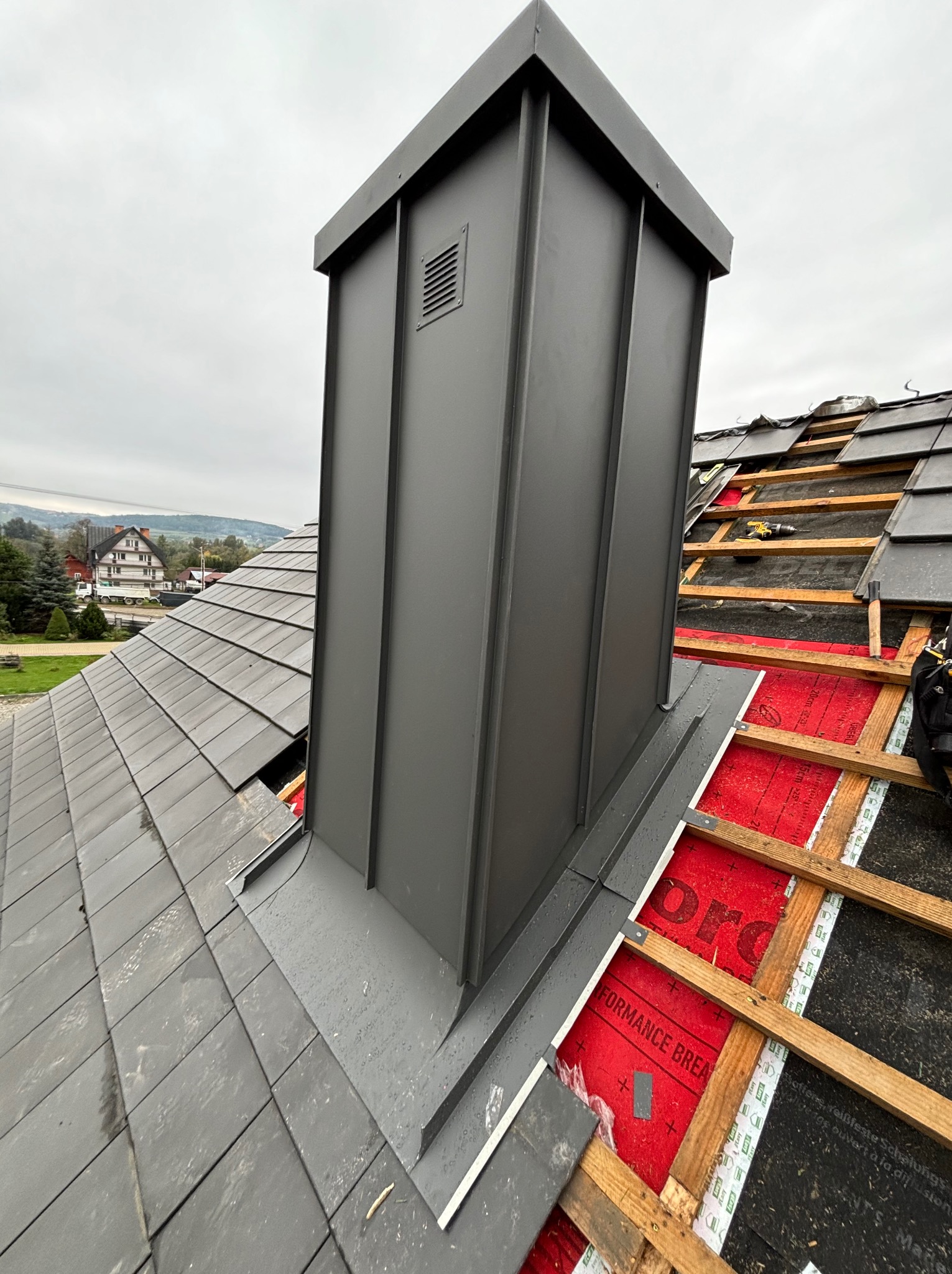 Nowy, szary komin wentylacyjny z blachy na dachu pokrytym szarą dachówką, częściowo zdjętą podczas prac. Widoczne drewniane łaty, membrana dachowa i narzędzia. W tle zabudowa jednorodzinna...