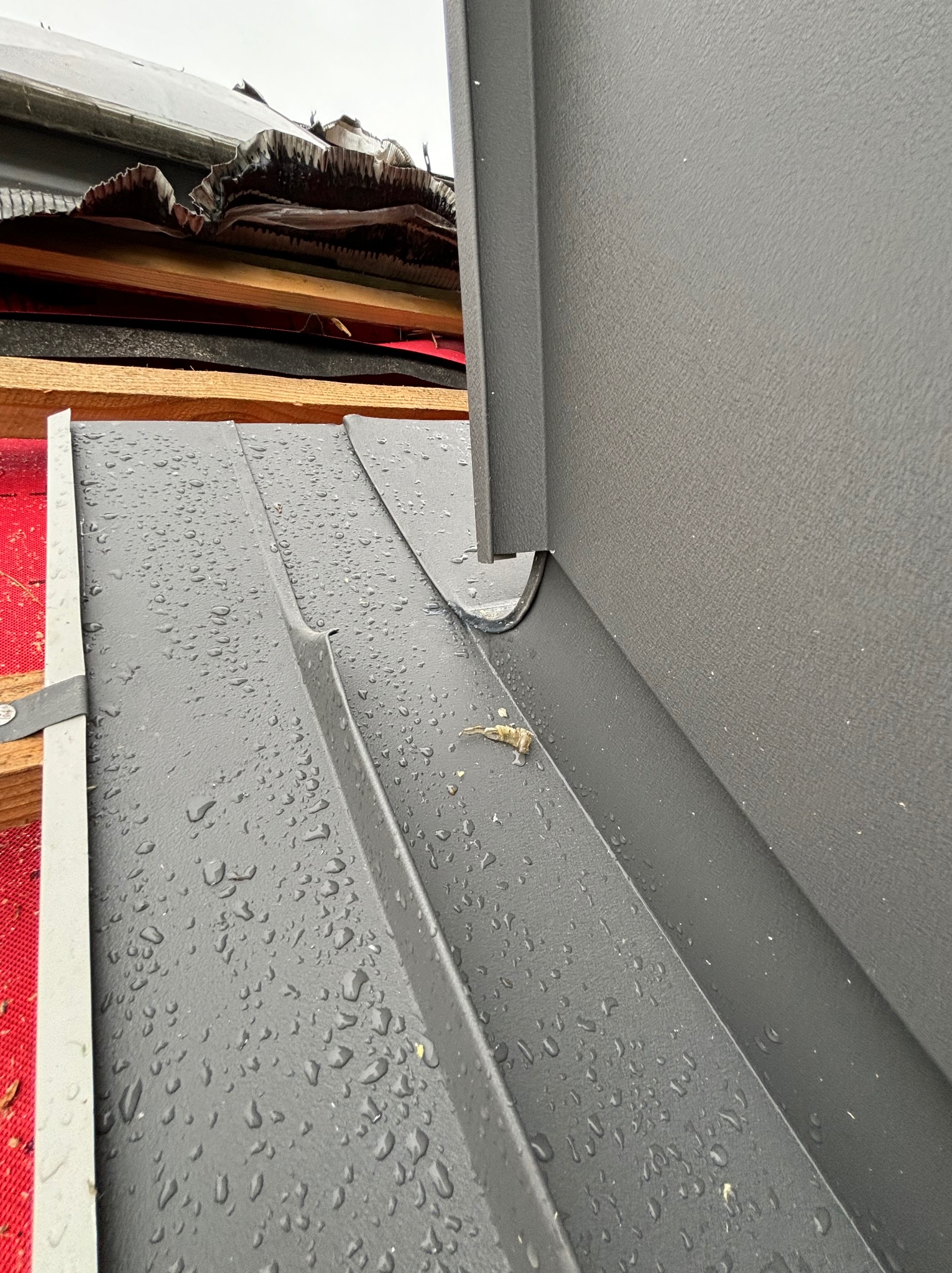 Szare, metalowe elementy dachu pokryte kroplami deszczu, widoczne drewniane elementy konstrukcji i fragment czerwonej membrany dachowej.