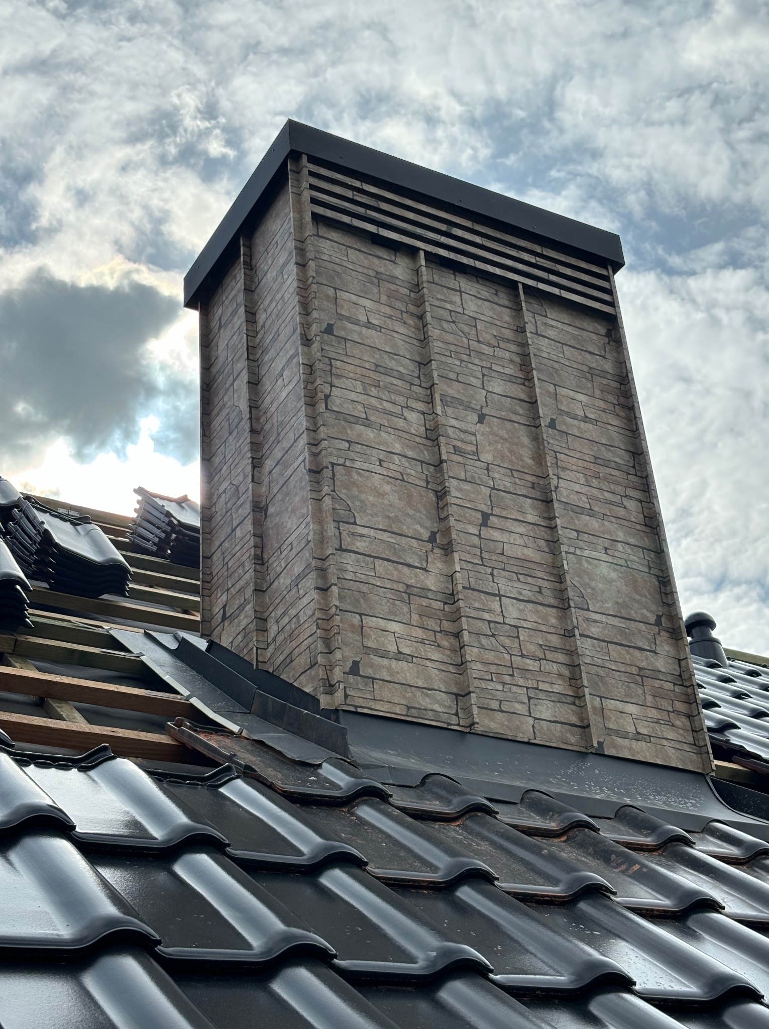 Montaż czarnej dachówki ceramicznej w Limanowej, widoczny komin obłożony imitacją kamienia i przygotowane drewniane łaty pod dach. Częściowo zachmurzone niebo w tle.