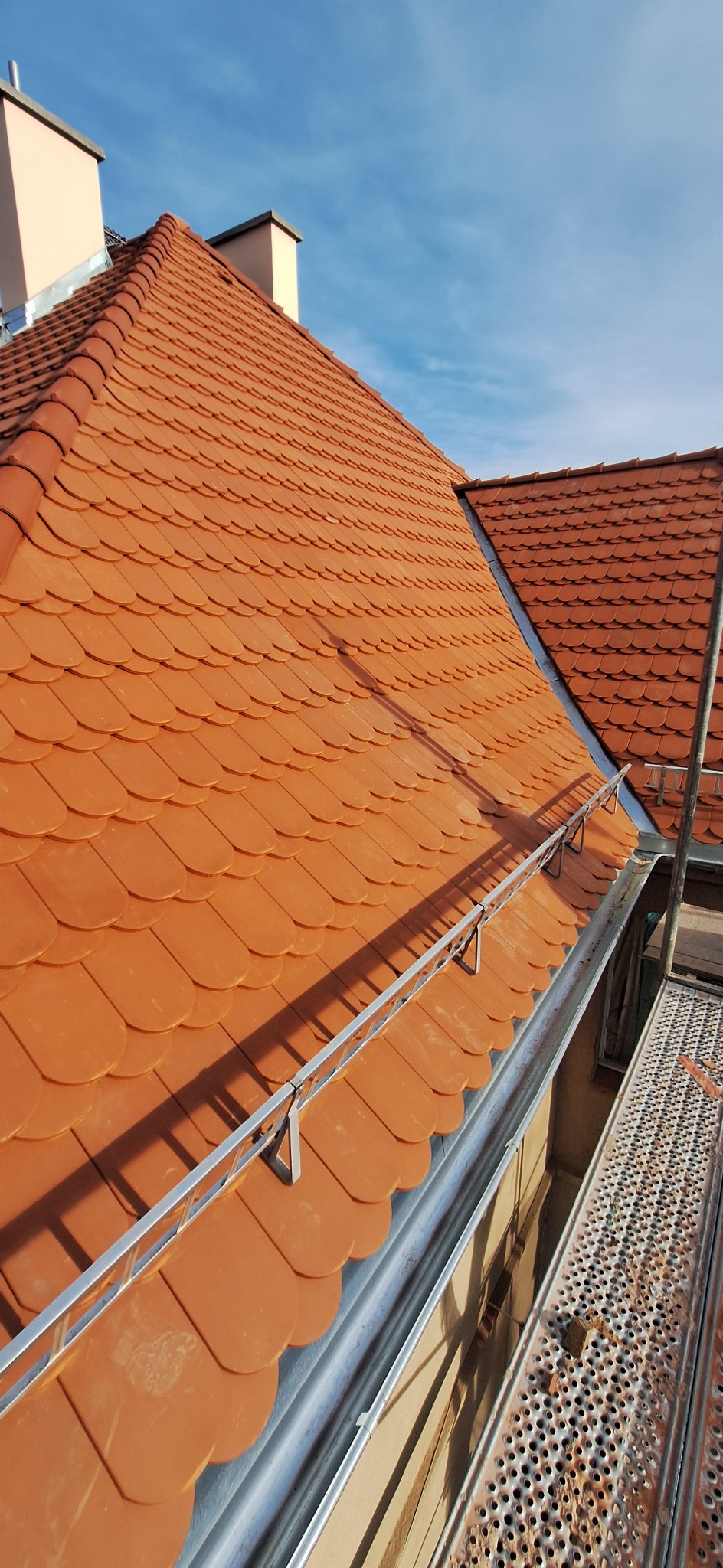 Czerwona dachówka karpiówka na dachu budynku, widok z rusztowania. Zamontowane zabezpieczenia przeciwśniegowe i rynna. Błękitne niebo w tle.