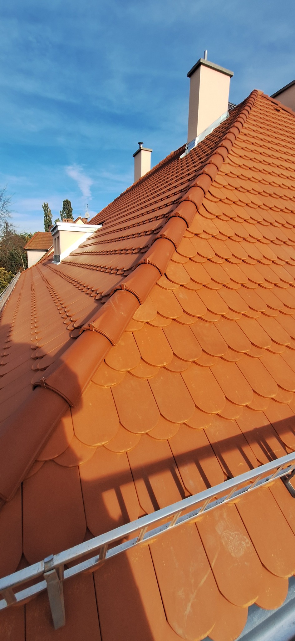 Dachówka karpiówka w odcieniach terakoty, widok z góry, z fragmentem drabiny dekarskiej. W tle kominy i błękitne niebo.