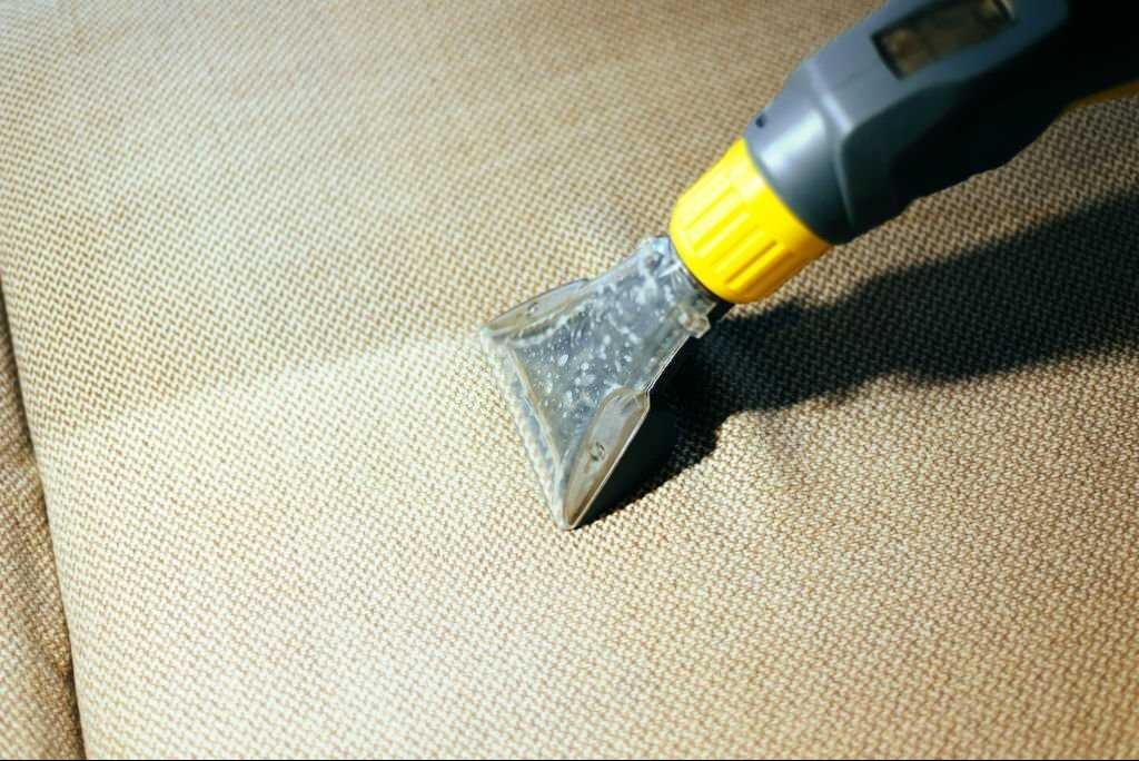 Czyszczenie beżowej tapicerki kanapy odkurzaczem piorącym z przezroczystą ssawką, widoczna różnica w kolorze po czyszczeniu.