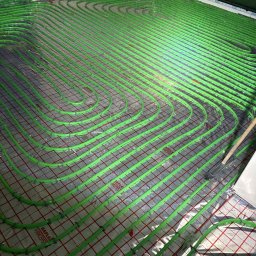 Bartosz Gładysz Termotechnik - Zielone rury ogrzewania podłogowego ułożone na siatce, blisko ściany z oknem. Widoczne mocowania rur i fragment narzędzia. Instalacja w trakcie montażu.