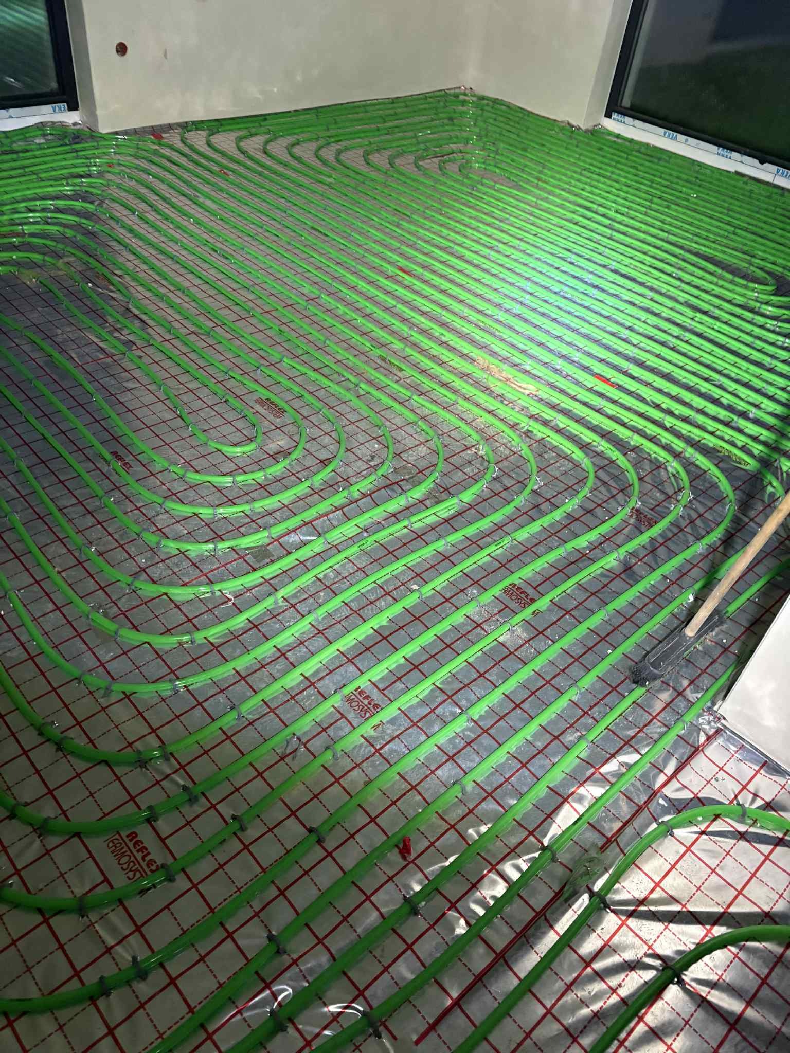 Zielone rury ogrzewania podłogowego ułożone na siatce, blisko ściany z oknem. Widoczne mocowania rur i fragment narzędzia. Instalacja w trakcie montażu.