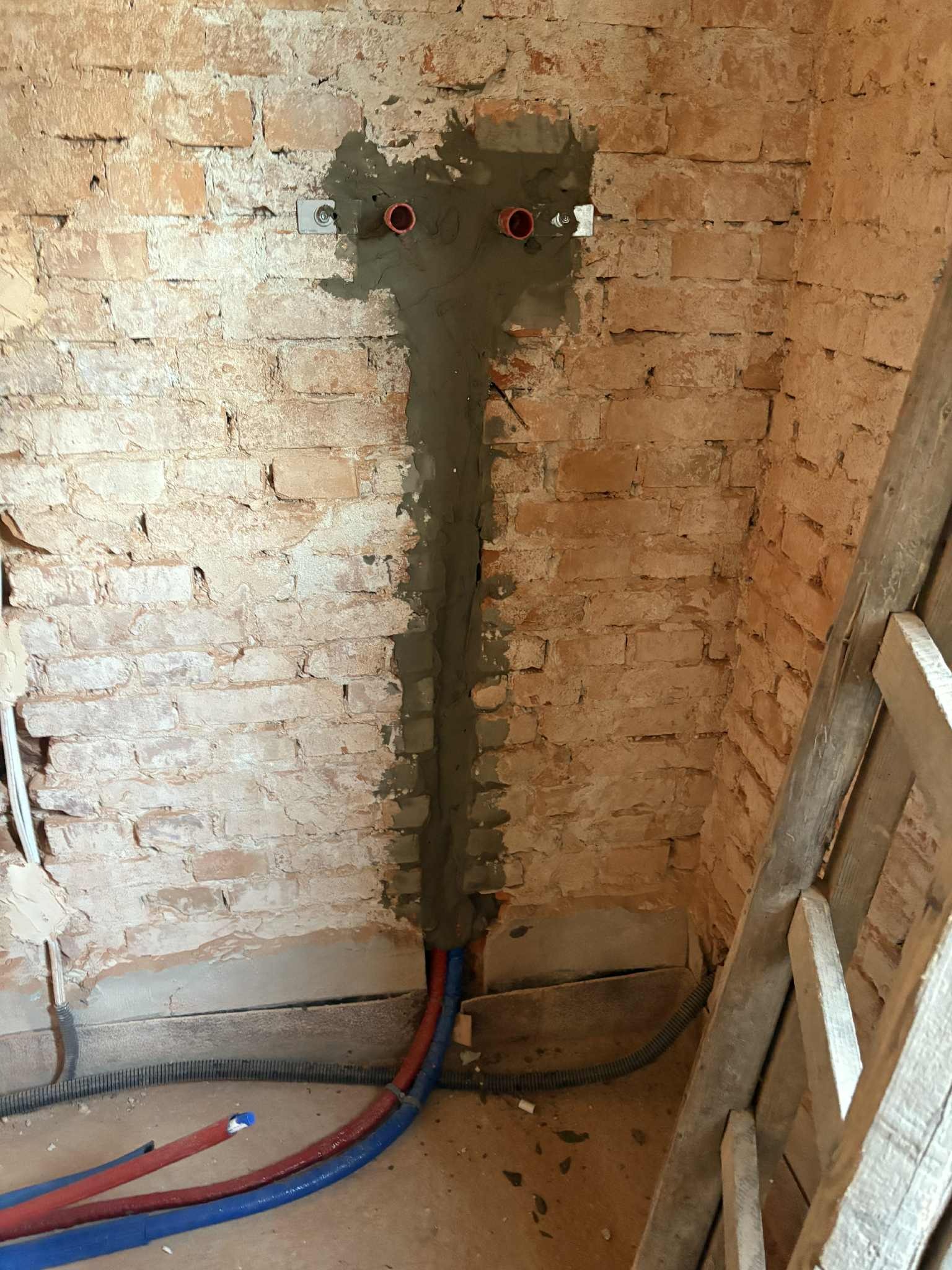 Czerwono-niebieskie rury instalacji wodnej w bruzdach ceglanej ściany, zamurowane szarą zaprawą. Widoczne gniazdka i fragment drabiny.