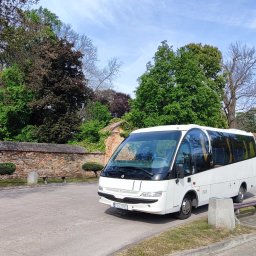 MIKA BUS Łukasz Mika - Biały minibus zaparkowany na tle kamiennego muru i zieleni, w tle widoczne budynki.