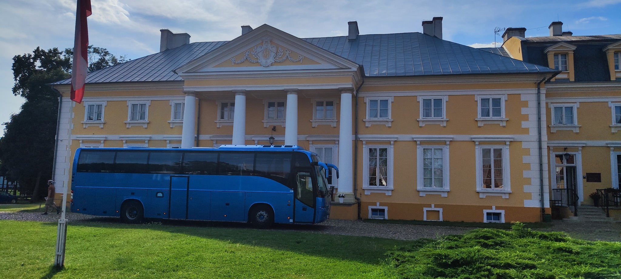 Niebieski autokar turystyczny zaparkowany przed żółtym pałacem z kolumnami w słoneczny dzień.