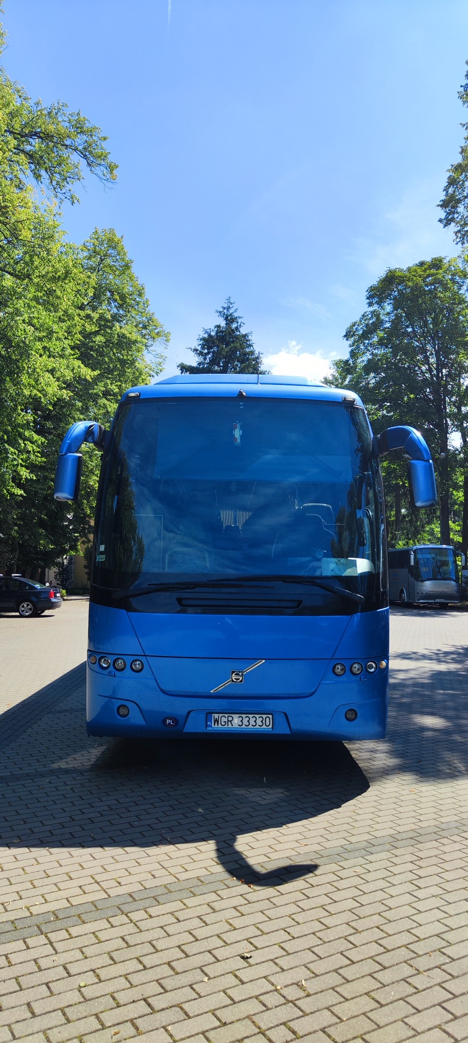 Niebieski autokar Volvo z polską rejestracją WGR 33330, zaparkowany na brukowanym placu w słoneczny dzień, z drzewami w tle.