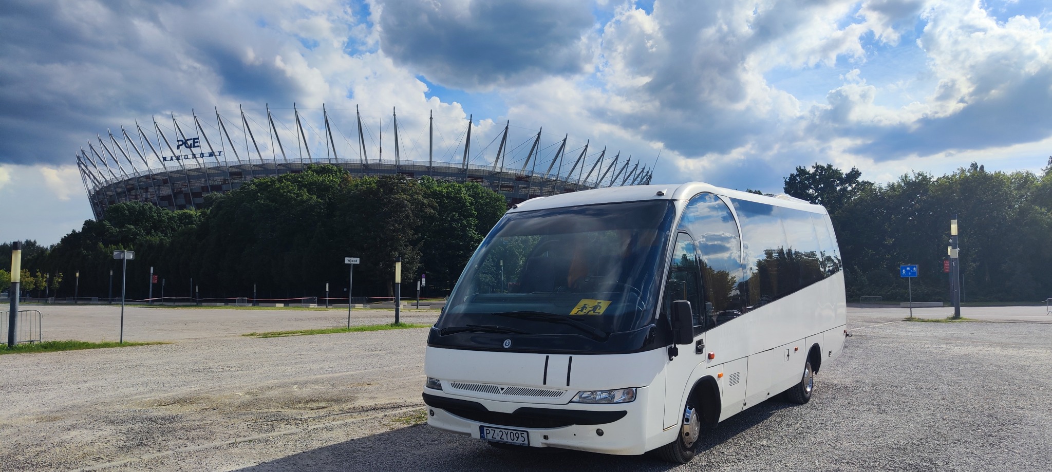 Biały bus z naklejką z sylwetkami dzieci na przedniej szybie, zaparkowany na tle Stadionu Narodowego w Warszawie.