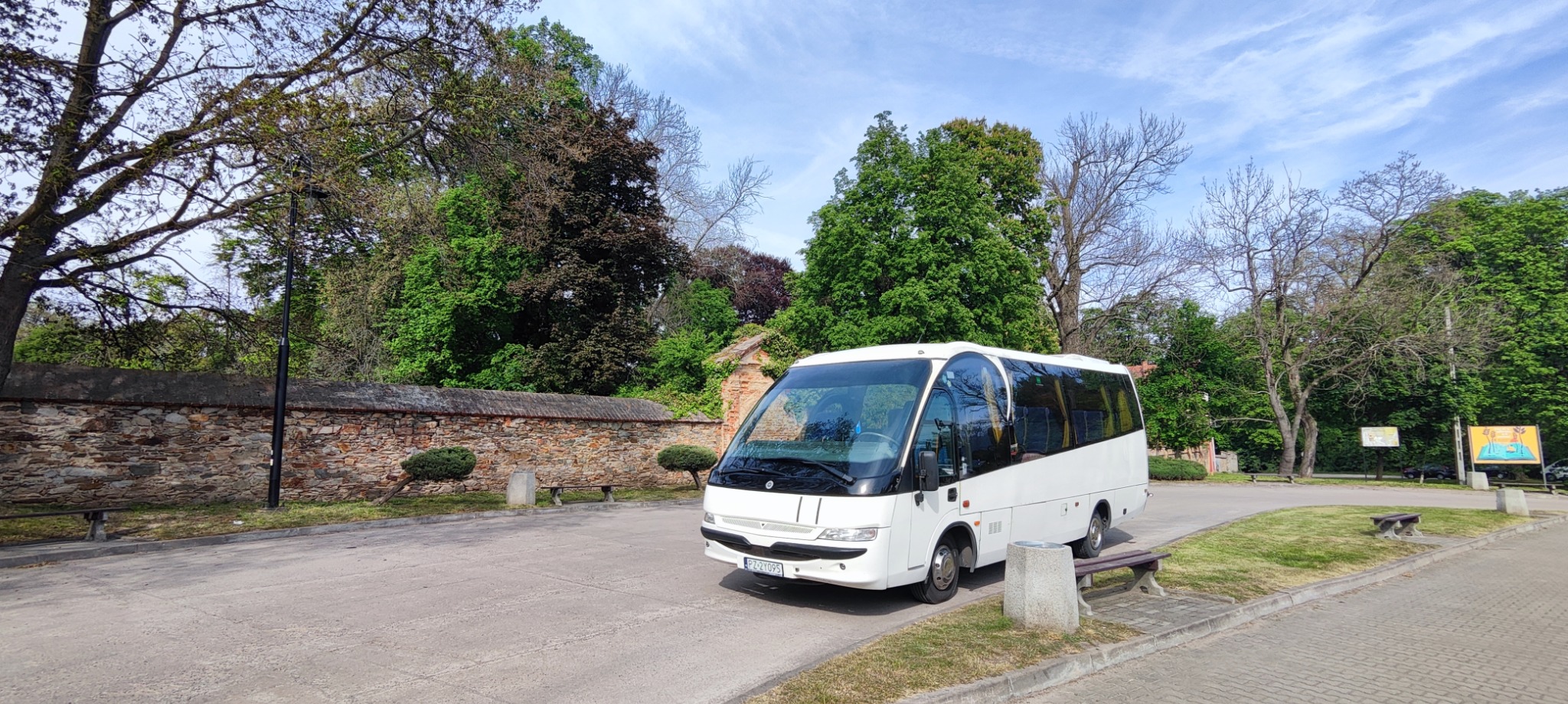 Biały minibus zaparkowany na tle kamiennego muru i zieleni, w tle widoczne budynki.