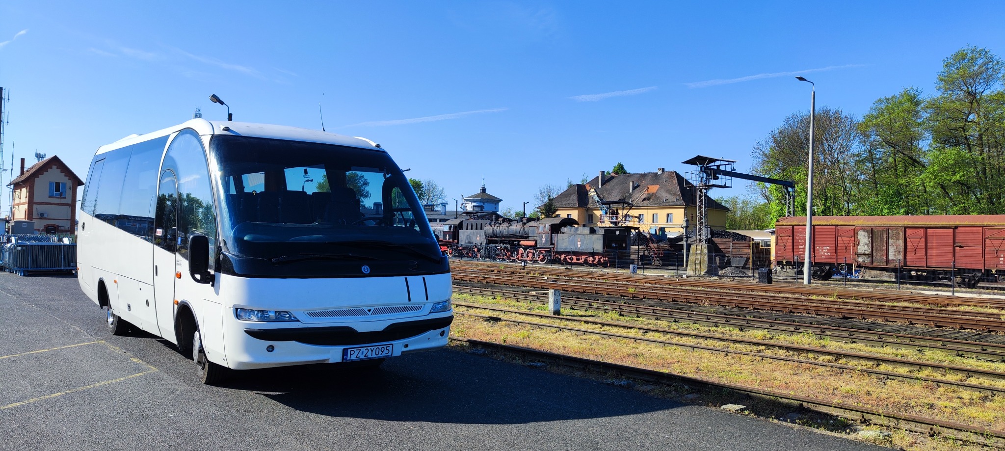 Biały bus pasażerski na tle skansenu kolejowego z lokomotywami i wagonami towarowymi w słoneczny dzień.