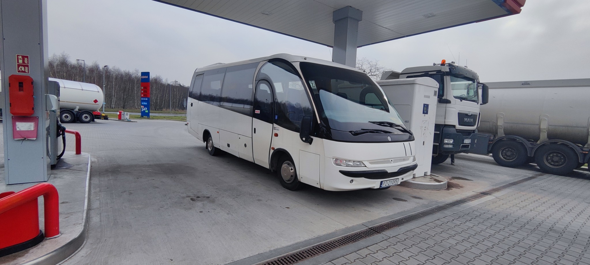 Biały bus pasażerski zaparkowany na stacji benzynowej obok dystrybutora i ciężarówki z cysterną.