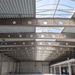 Wnętrze hali przemysłowej z widoczną konstrukcją dachu, betonowymi belkami z okrągłymi otworami i zamontowanym oświetleniem punktowym.