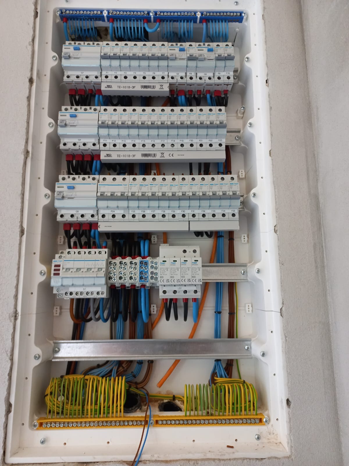 Rozdzielnica elektryczna w białej obudowie, widoczne zabezpieczenia nadprądowe, przewody w kolorach niebieskim, brązowym, czarnym i żółto-zielonym. Dolna część rozdzielnicy z listwą zaciskową...