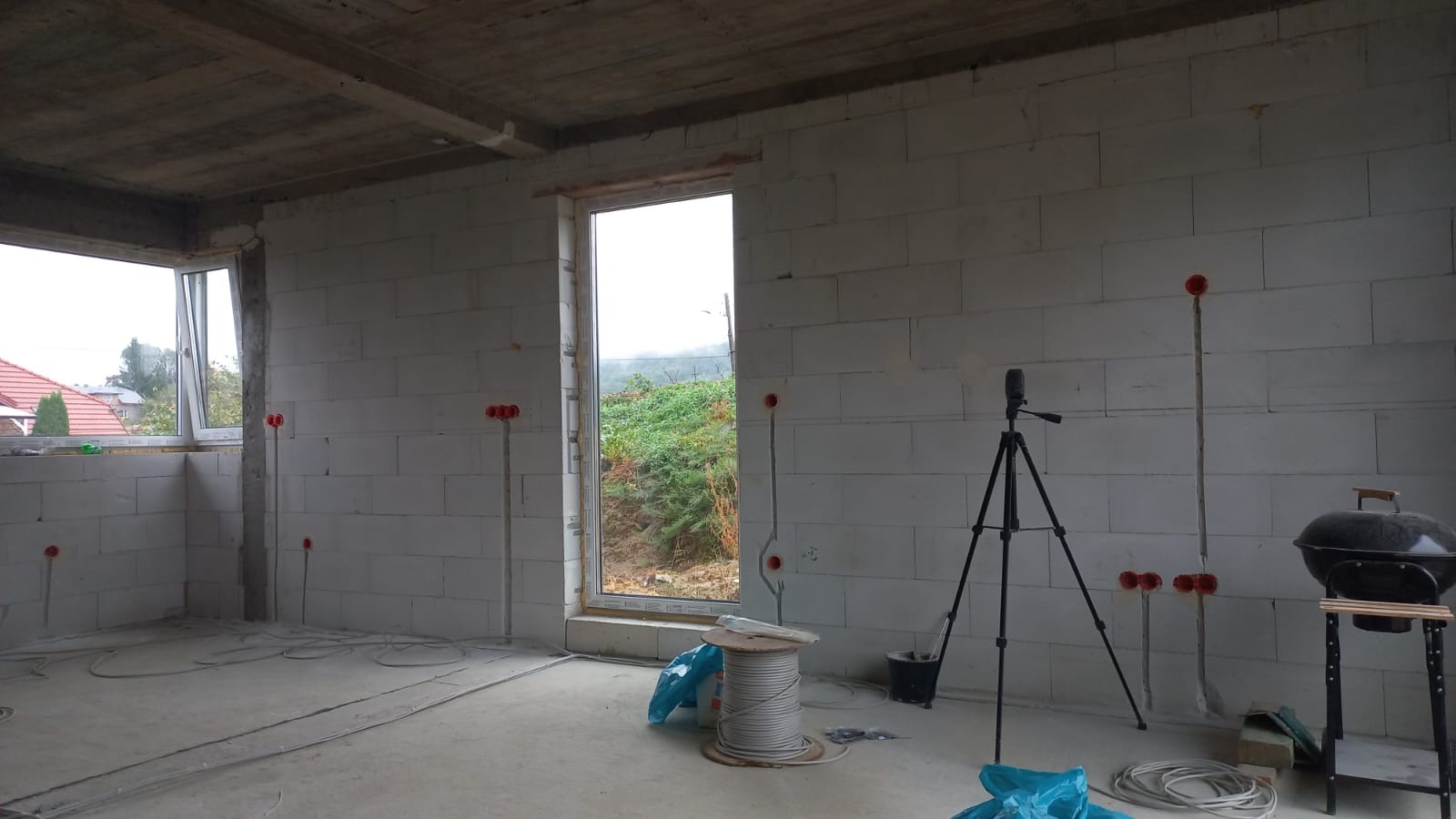 Wnętrze surowego domu w trakcie instalacji elektrycznej: widoczne przewody, puszki elektryczne, rolka kabla, poziomica na statywie i grill na kółkach.
