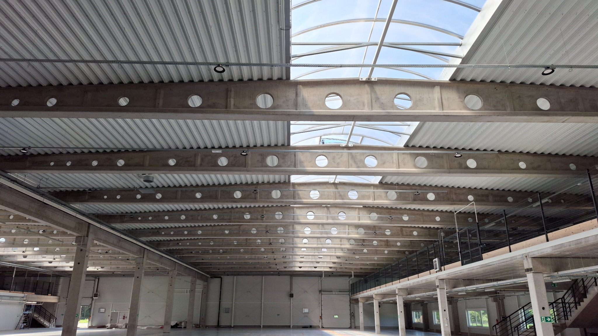 Wnętrze hali przemysłowej z widoczną konstrukcją dachu, betonowymi belkami z okrągłymi otworami i zamontowanym oświetleniem punktowym.