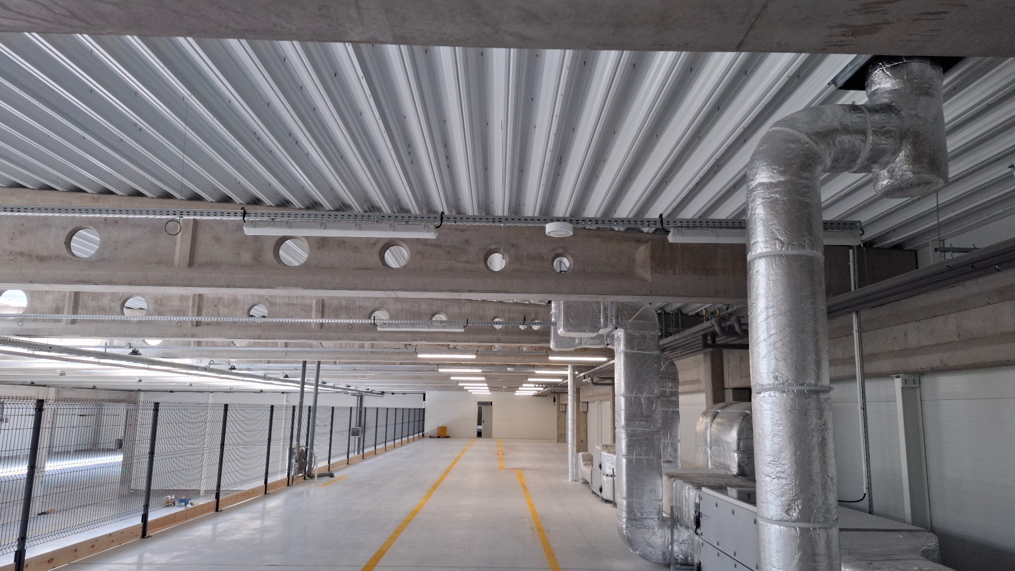 Wnętrze hali produkcyjnej z instalacją oświetleniową: betonowe belki z okrągłymi otworami i zamontowanymi lampami. Widoczne koryta kablowe i rury wentylacyjne.