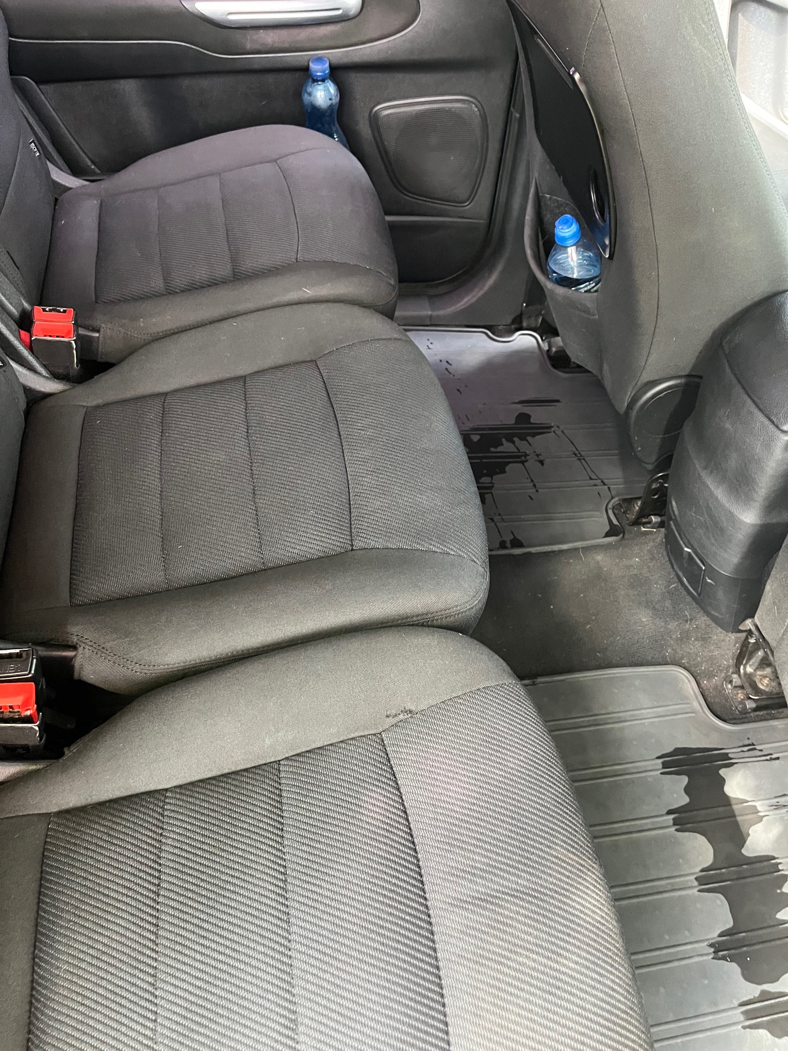 Wnętrze samochodu z czarną tapicerką siedzeń po czyszczeniu, widoczne mokre ślady na gumowych dywanikach i butelka wody w kieszeni fotela.