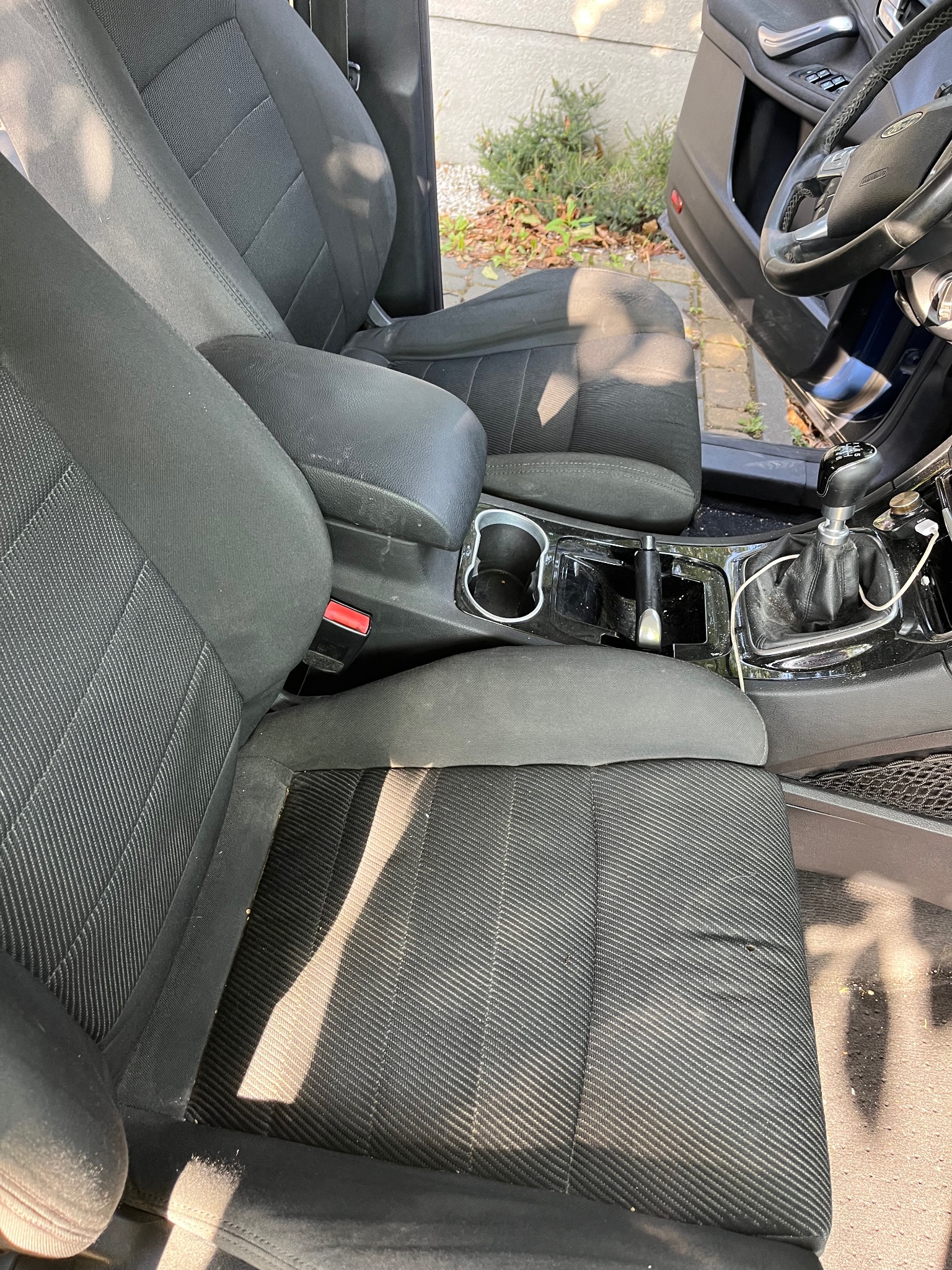 Wnętrze samochodu przed czyszczeniem: widoczne zabrudzenia na czarnej tapicerce foteli, podłokietniku i konsoli środkowej, zarysowane elementy plastikowe.