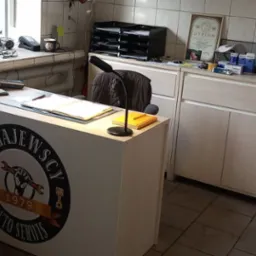 Recepcja warsztatu samochodowego z logo 'ZAGAJEWSCY AUTO SERWIS 1979', na blacie biurka leżą dokumenty i lampka, w tle regał z dyplomem i akcesoriami.