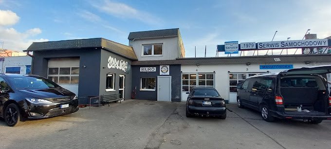 Zewnętrzny widok warsztatu samochodowego z trzema stanowiskami, zaparkowanymi samochodami osobowymi i napisem 'Serwis Samochodowy' na dachu budynku.