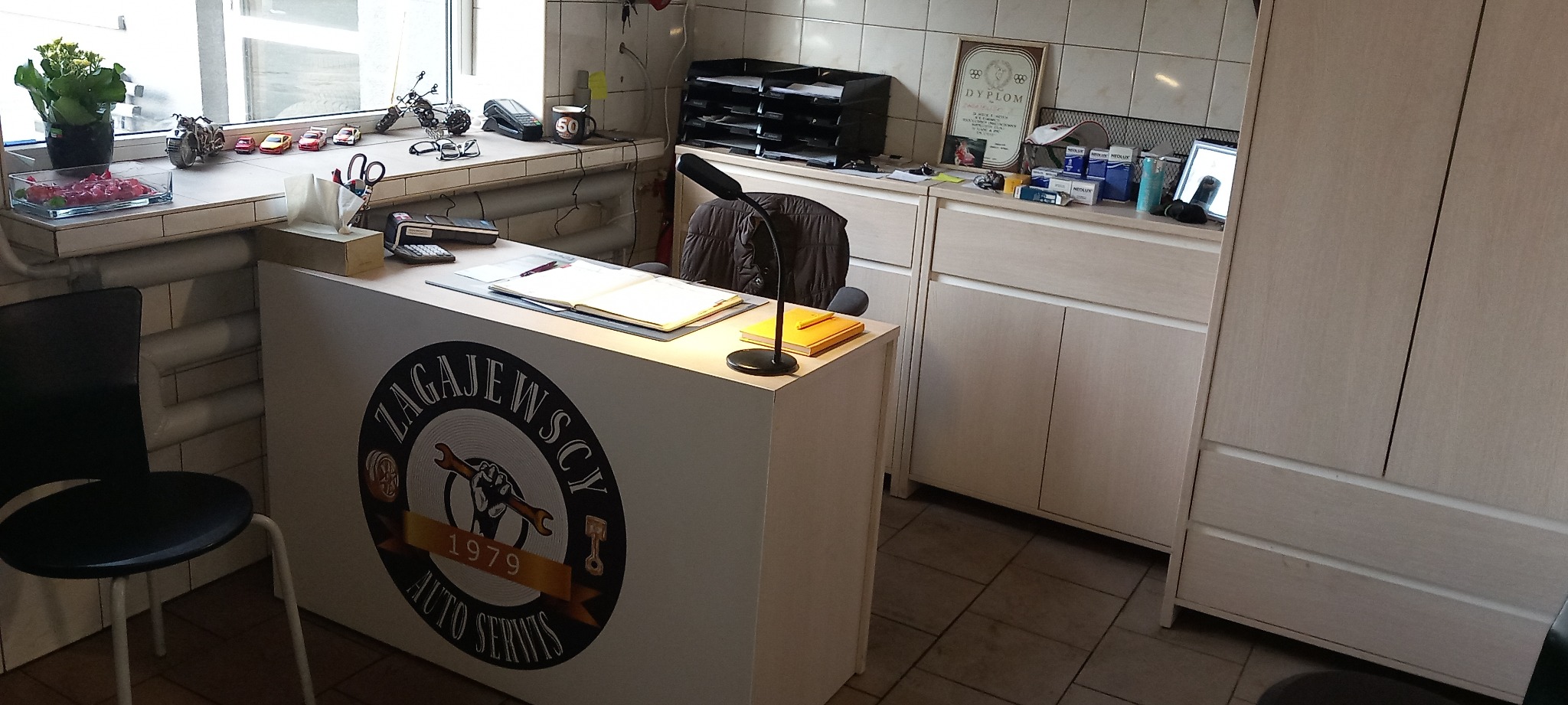 Recepcja warsztatu samochodowego z logo 'ZAGAJEWSCY AUTO SERWIS 1979', na blacie biurka leżą dokumenty i lampka, w tle regał z dyplomem i akcesoriami.