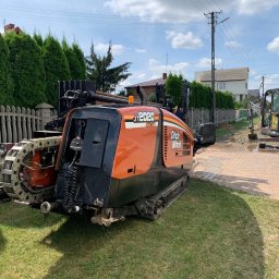 ABDRILL SPÓŁKA Z OGRANICZONĄ ODPOWIEDZIALNOŚCIĄ - Pomarańczowo-czarna maszyna Ditch Witch JT2020 Mach1 do przewiertów sterowanych na trawniku, w tle wykop pod instalacje wykonywany małą koparką Volvo.
