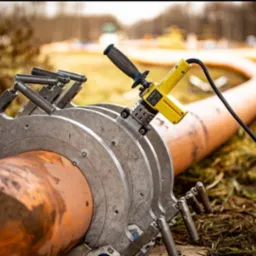 Zgrzewarka doczołowa do rur z tworzyw sztucznych, widoczna w trakcie pracy na zewnątrz, rura lekko zabrudzona ziemią.