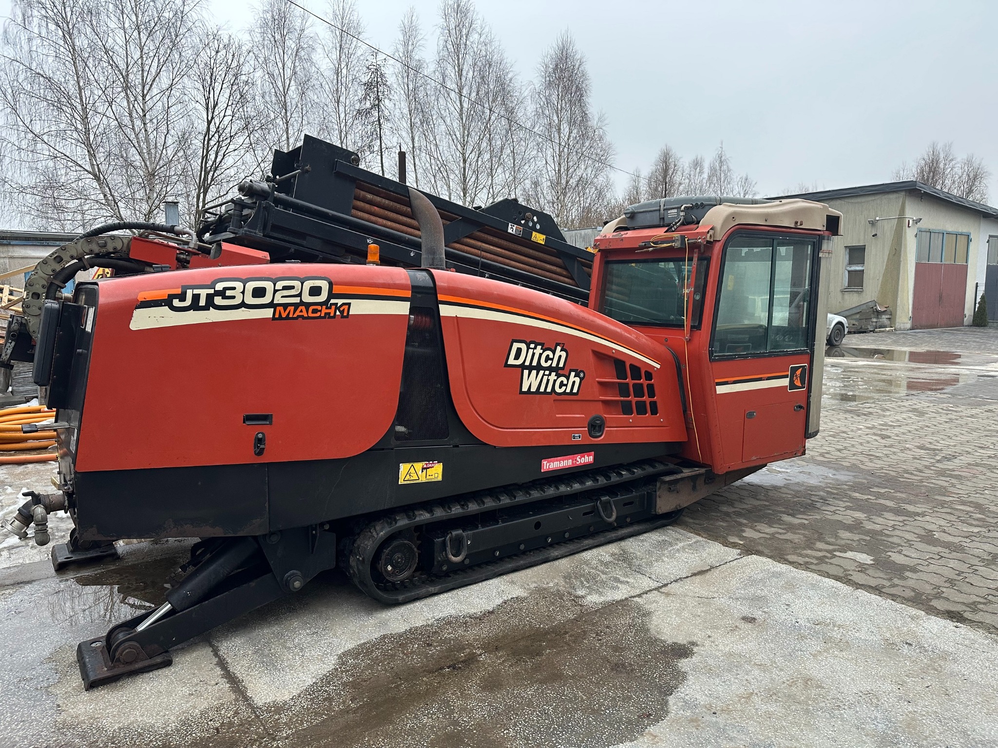 Pomarańczowa maszyna Ditch Witch JT3020 Mach 1 na gąsienicach, z widocznymi rurami w tle i budynkiem w oddali, stojąca na betonowej nawierzchni.