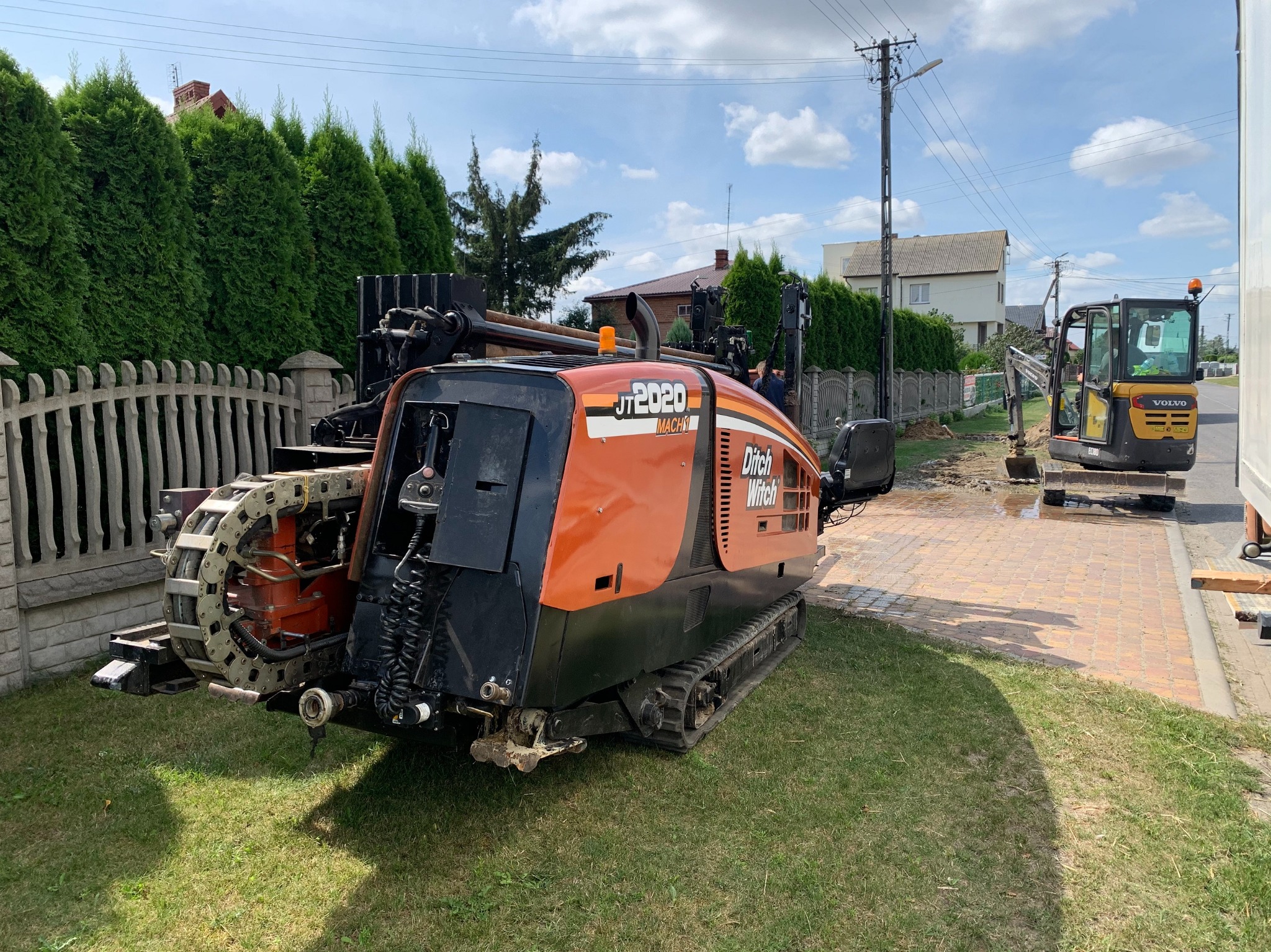Pomarańczowo-czarna maszyna Ditch Witch JT2020 Mach1 do przewiertów sterowanych na trawniku, w tle wykop pod instalacje wykonywany małą koparką Volvo.