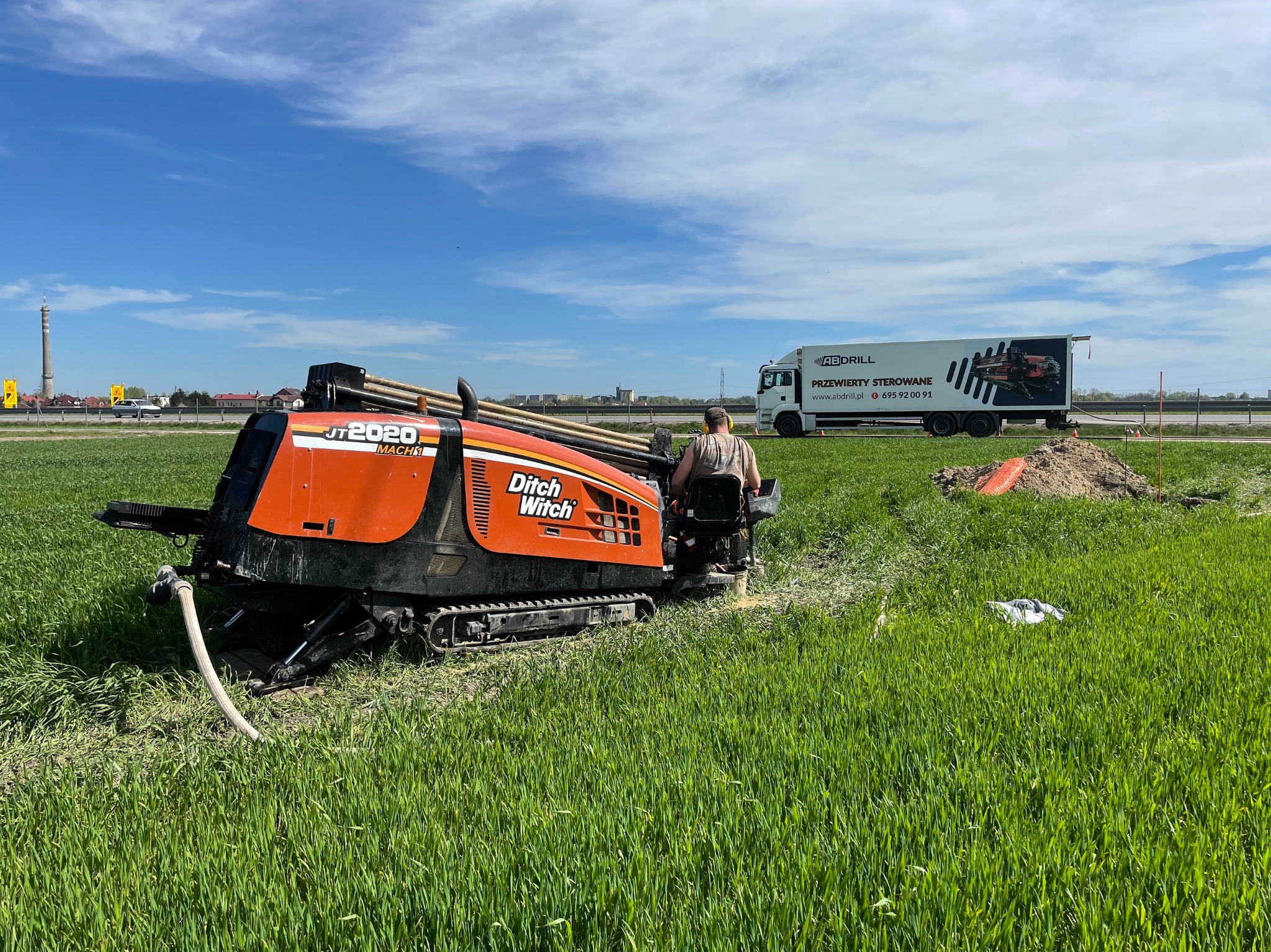 Pomarańczowa maszyna Ditch Witch JT2020 Mach1, operator w kabinie, pracuje na polu z zieloną trawą, w tle biała ciężarówka z logo firmy AB Drill i hasłem 'Przewierty Sterowane'.