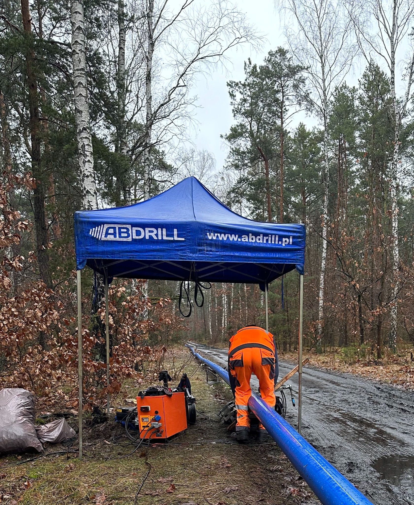 Pracownik w pomarańczowym kombinezonie łączy rury na leśnej drodze, obok stoi pomarańczowa maszyna i namiot z logo firmy AB Drill.