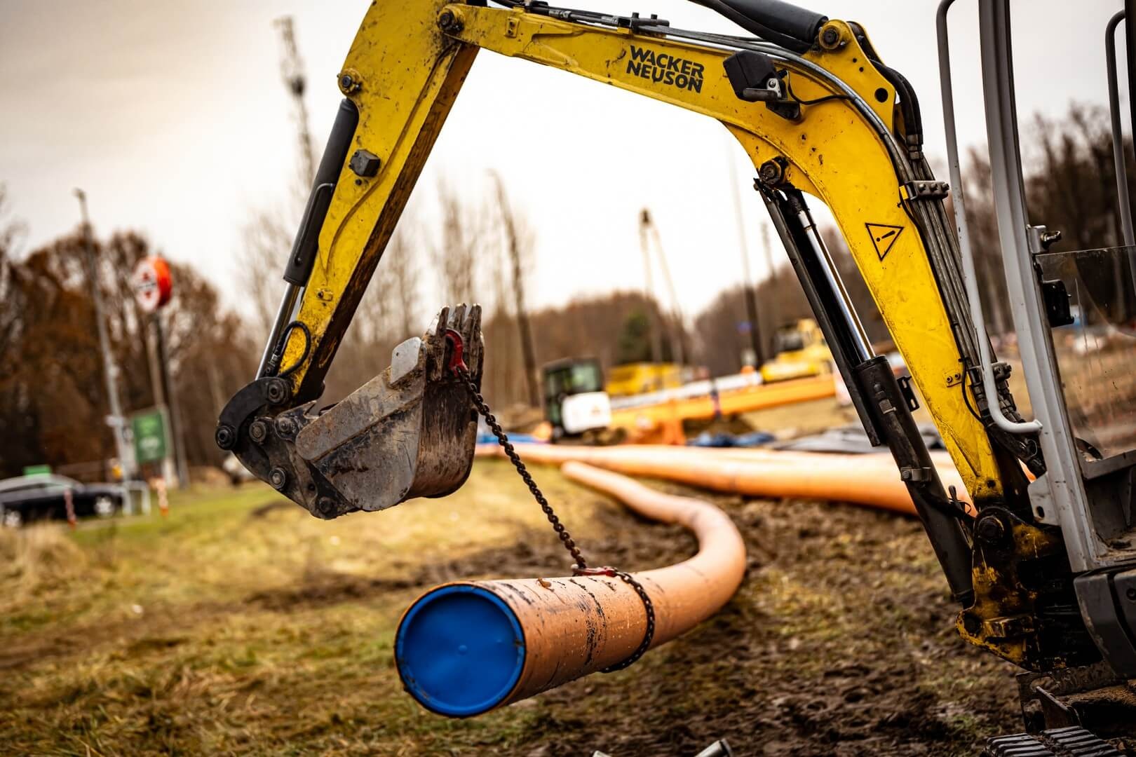 Żółta koparka Wacker Neuson w Warszawie podnosi rurę z niebieskim zakończeniem za pomocą łańcucha, w tle widoczne inne rury i maszyny budowlane.
