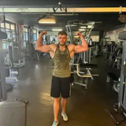 Mężczyzna z brodą, w sportowej koszulce na ramiączkach i krótkich spodenkach, prezentuje swoją muskulaturę w siłowni, z widocznymi maszynami do ćwiczeń w tle.
