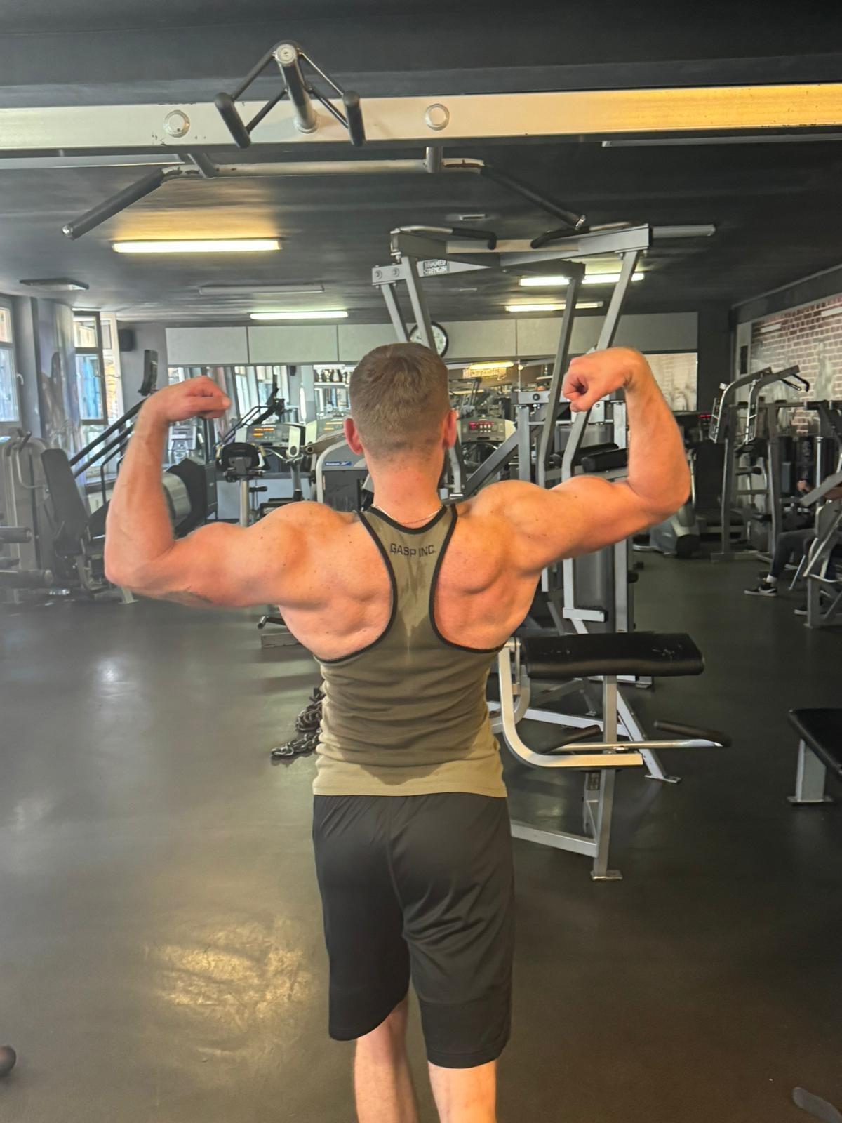 Mężczyzna w sportowej koszulce, odwrócony tyłem, prezentuje umięśnione plecy i bicepsy na tle siłowni z maszynami do ćwiczeń.