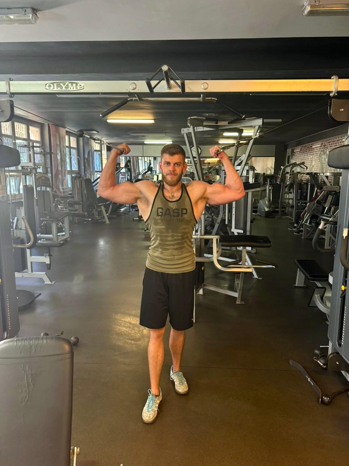 Mężczyzna z brodą, w sportowej koszulce na ramiączkach i krótkich spodenkach, prezentuje swoją muskulaturę w siłowni, z widocznymi maszynami do ćwiczeń w tle.
