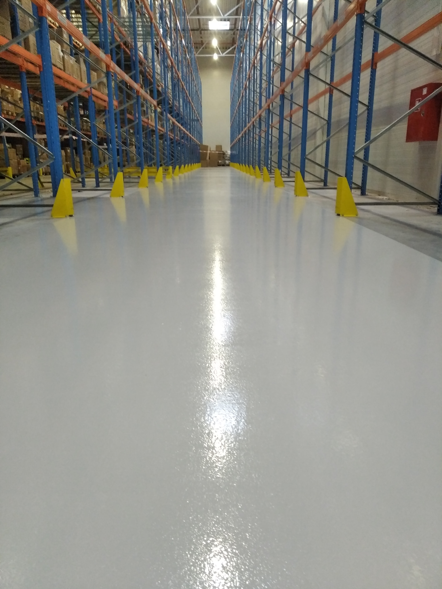 Błyszcząca, jasnoszara posadzka żywiczna w magazynie, z żółtymi osłonami słupów i regałami w tle.