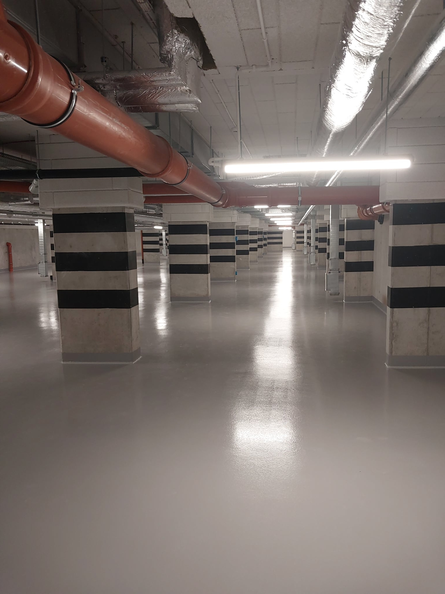 Jasnoszara, gładka powierzchnia w garażu podziemnym z rzędami kolumn w czarno-białe pasy i pomarańczowymi rurami pod sufitem.