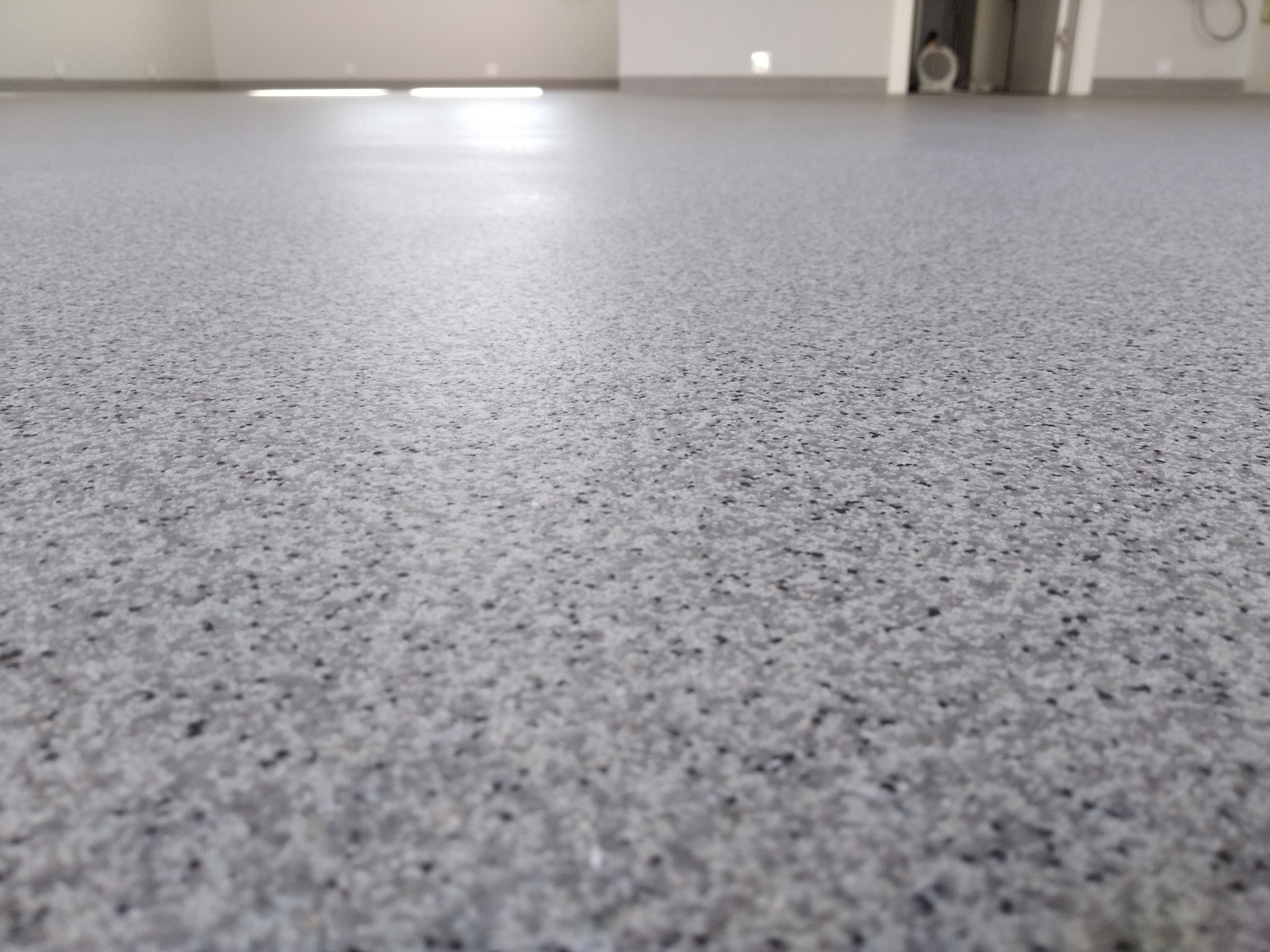 Szara, gładka podłoga z drobnymi, czarnymi plamkami, widziana z niskiej perspektywy w jasnym, pustym pomieszczeniu. W tle widoczne białe ściany i drzwi.