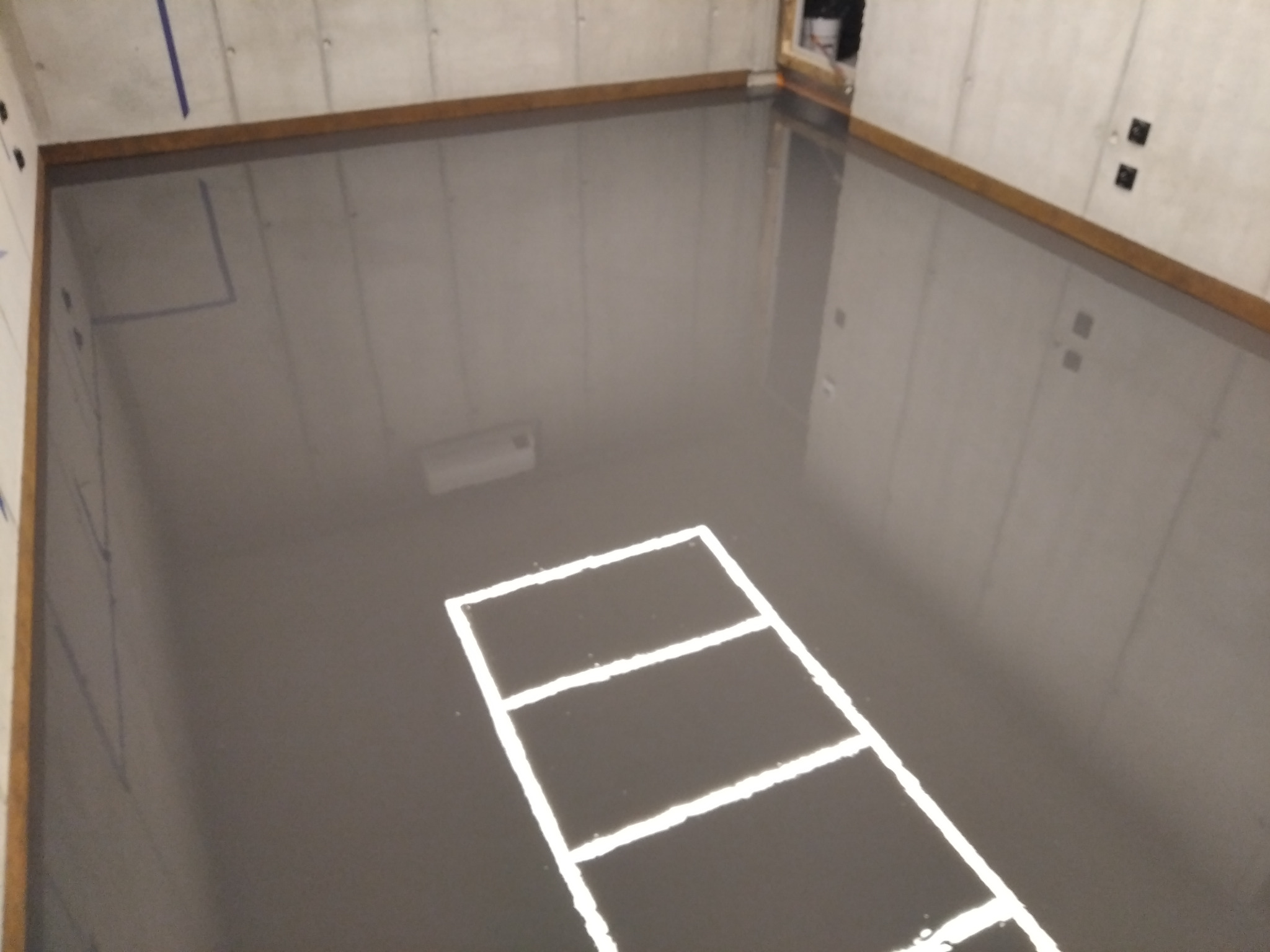 Szara, błyszcząca posadzka w surowym pomieszczeniu z betonowymi ścianami i białymi liniami wyznaczającymi prostokątne pola.