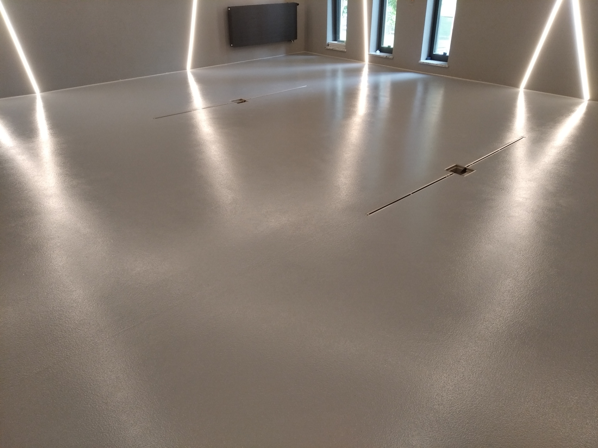 Gładka, szara podłoga z odpływami liniowymi w nowocześnie oświetlonym wnętrzu z grzejnikiem na ścianie i oknami w tle.