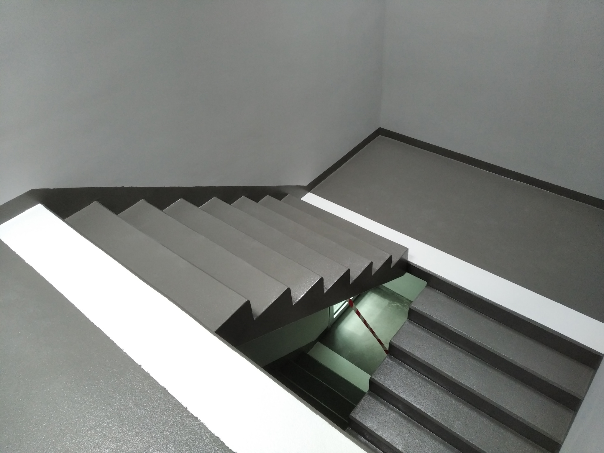 Klatka schodowa z szarymi schodami i białymi pasami na ścianach, widok z góry na dół.