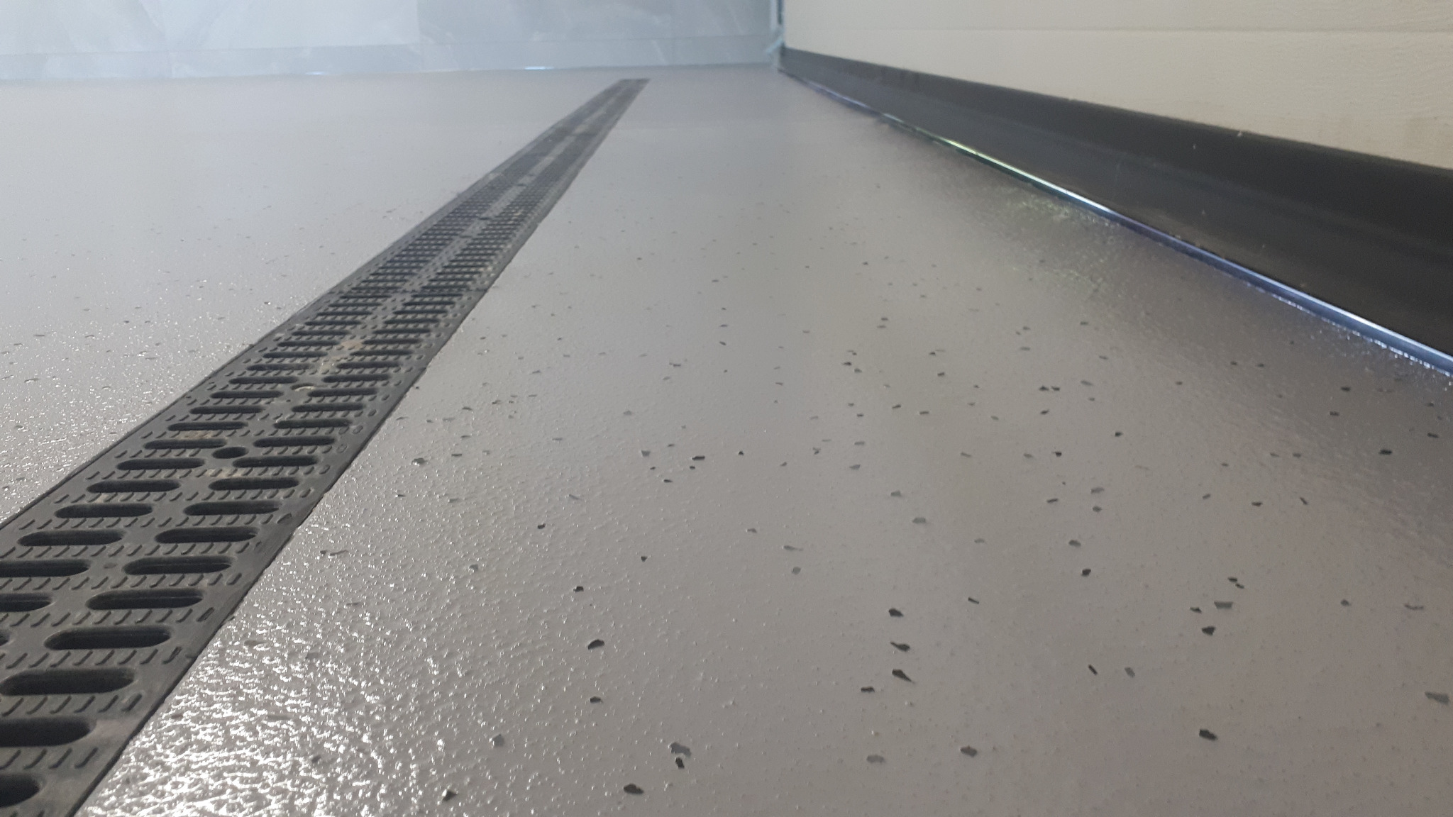 Szara, gładka posadzka z odpływem liniowym, widoczne drobne, ciemne wtrącenia w strukturze, listwa przypodłogowa w tle.