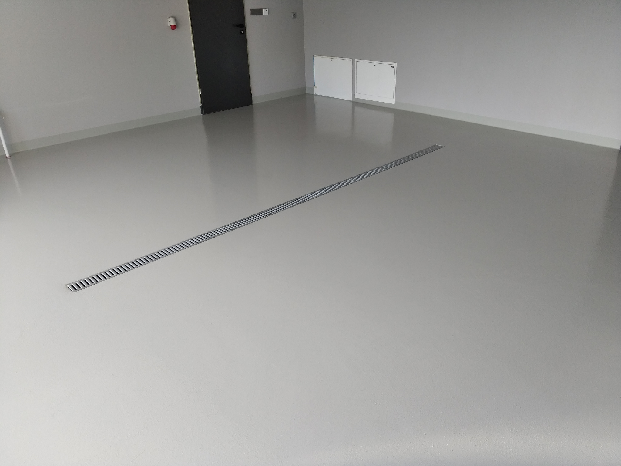 Jasnoszara, gładka podłoga z odpływem liniowym wzdłuż pomieszczenia, widoczne drzwi i białe skrzynki na ścianie.